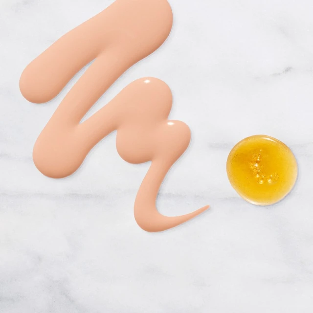 Swatches of skin-toned makeup foundation and yellow serum on a marble surface show product texture and color for beauty applications.