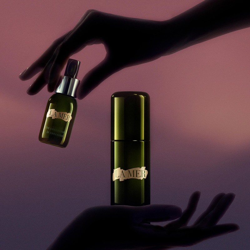 La Mer skincare bottles are held in the air by silhouetted hands against a lavender-pink gradient background, showcasing beauty product arrangement.