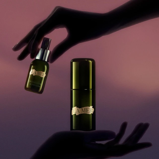 La Mer skincare bottles are held in the air by silhouetted hands against a lavender-pink gradient background, showcasing beauty product arrangement.