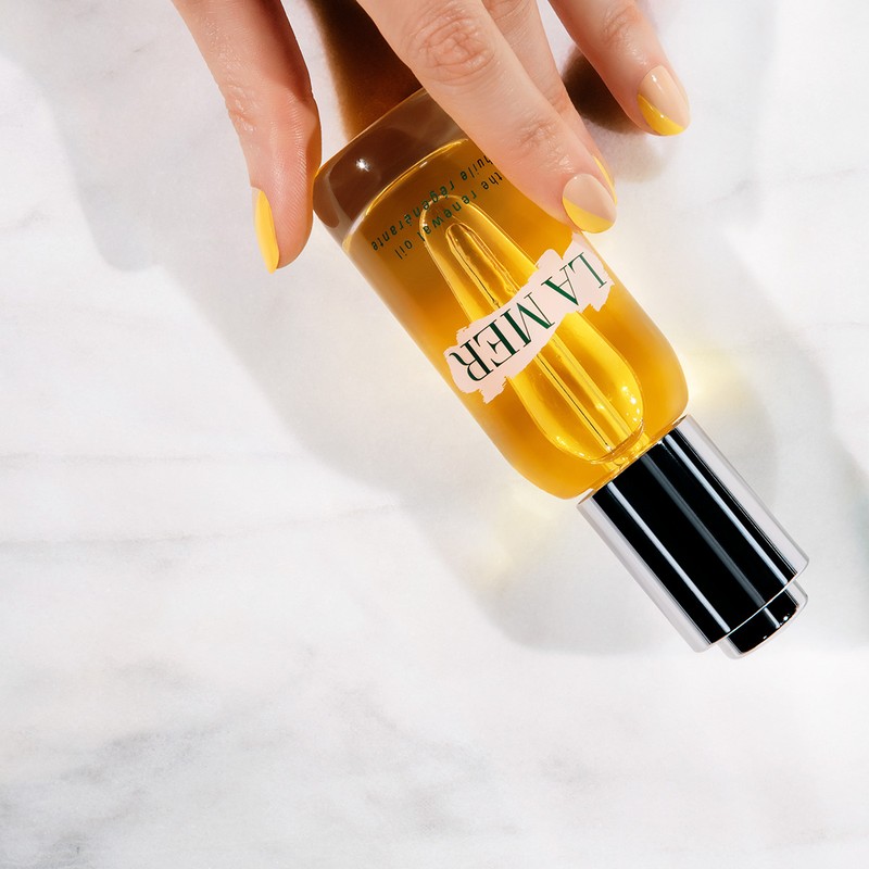 Close-up shot of La Mer renewal oil bottle with hand with yellow nail polish, set on a white marble background.