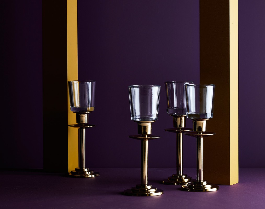 Still life shot of four clear glass candle holders with gold metal stands, set against a purple background with gold vertical stripes.