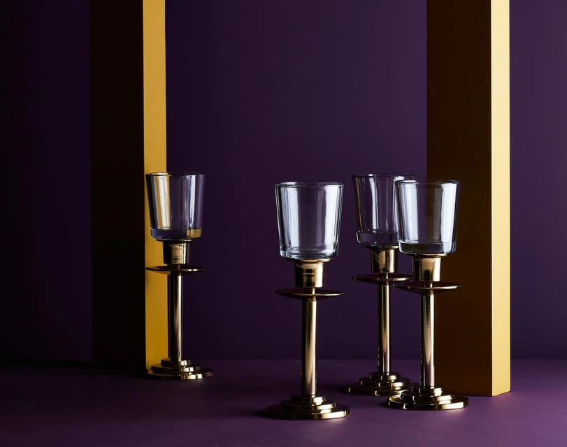 Still life shot of four clear glass candle holders with gold metal stands, set against a purple background with gold vertical stripes.