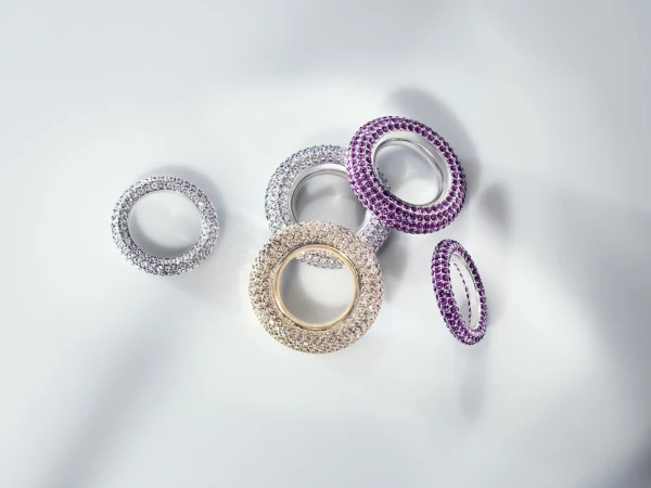 Several jeweled rings in silver, gold, and purple tones are arranged on a gradient white backdrop with soft, diffused lighting.