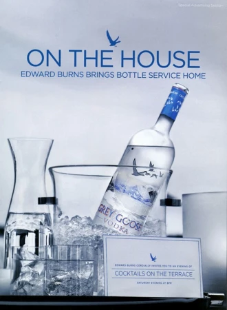 Grey Goose vodka bottle chilling in an ice bucket with glassware and a framed invitation card on a tray.