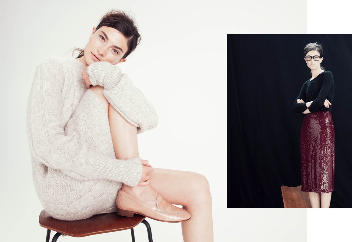 Two women in a fashion setting. One sits wearing a sweater and loafers, the other stands with crossed arms in a skirt and top.