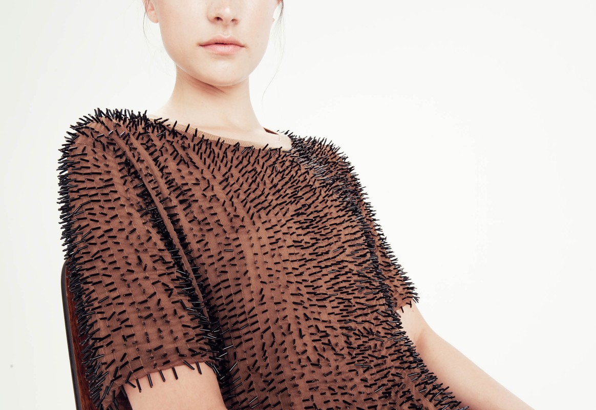 Fashion model wearing brown t-shirt with black pins in a studio setting on a white background in a medium shot with eye-level angle.