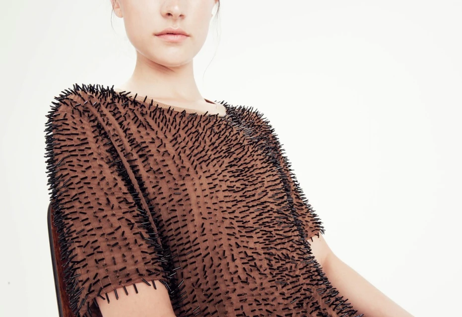 Fashion model wearing brown t-shirt with black pins in a studio setting on a white background in a medium shot with eye-level angle.