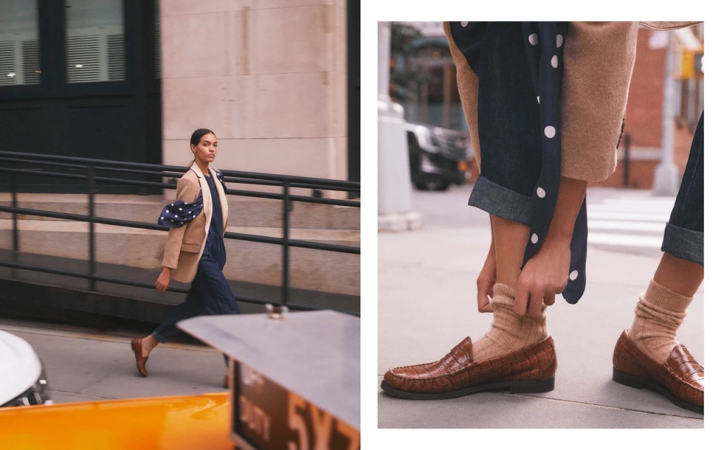 A stylish woman in a tan blazer walks in an urban setting near a yellow taxi. Close-up on brown loafer shoes with beige socks and rolled denim.
