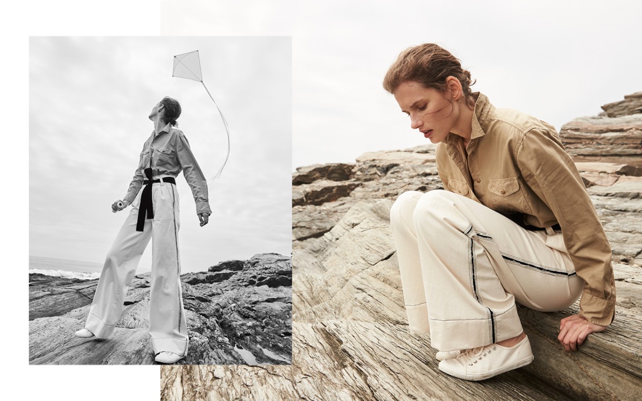 Fashion shot on rocky coast with woman in khaki shirt and white pants flying a kite. Neutral palette, natural light, editorial style.