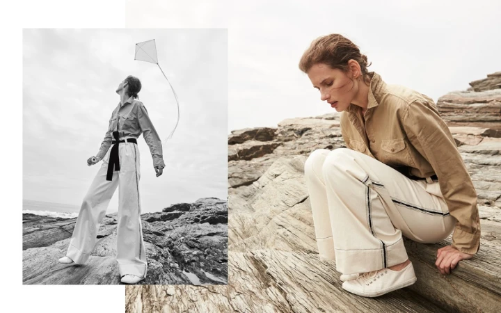 Fashion shot on rocky coast with woman in khaki shirt and white pants flying a kite. Neutral palette, natural light, editorial style.