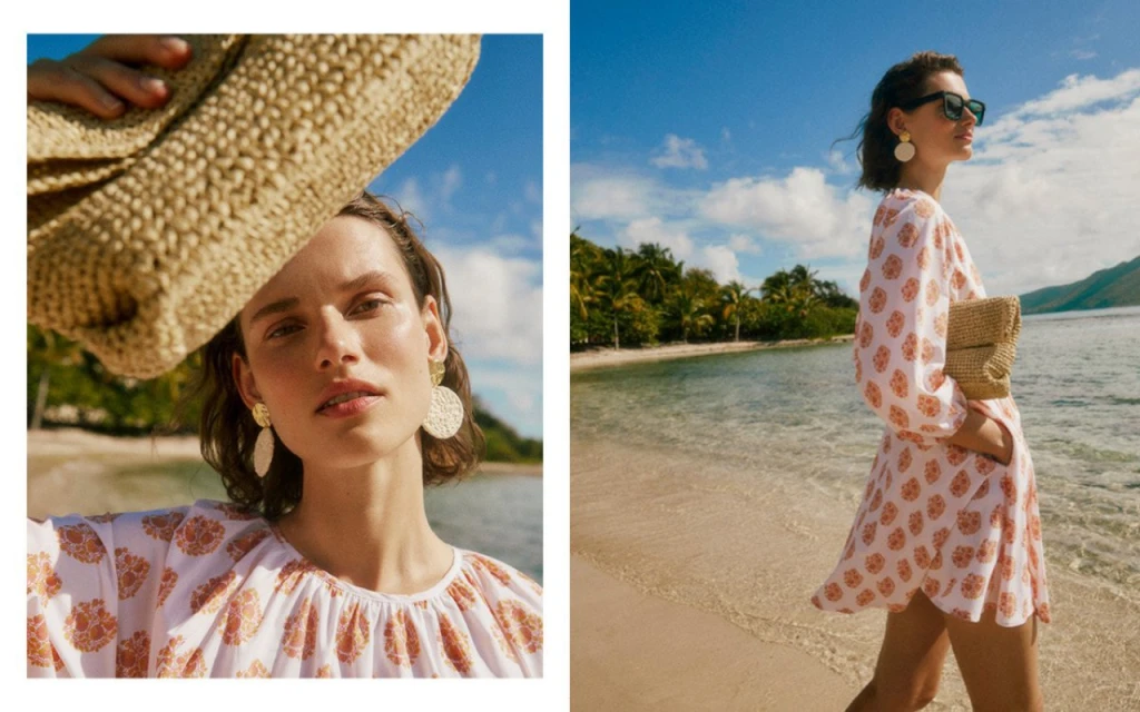 Woman on tropical beach with patterned dress and straw clutch bag. Summer fashion editorial shot with blue sky and palm trees.