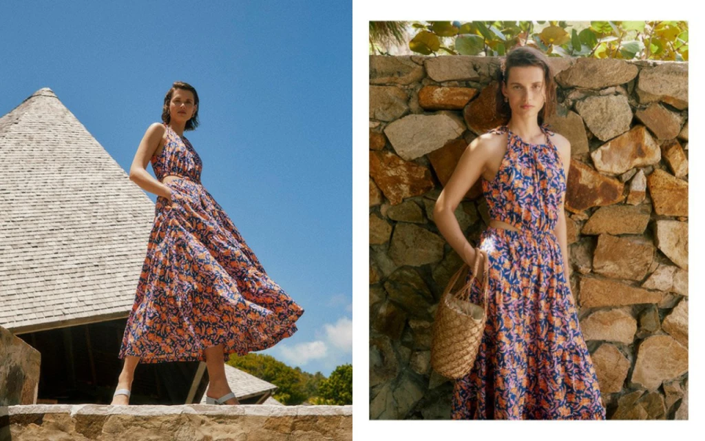 A woman in a flowing floral halter dress poses outdoors, featuring a cut-out waist and a straw bag, set against a stone wall and blue sky.