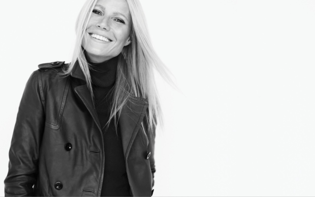 Monochrome portrait of Gwyneth Paltrow wearing a dark leather trench coat and turtleneck against a white background