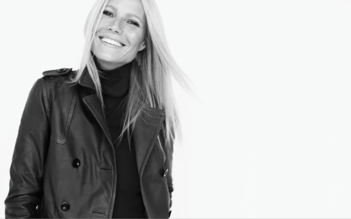 Monochrome portrait of Gwyneth Paltrow wearing a dark leather trench coat and turtleneck against a white background