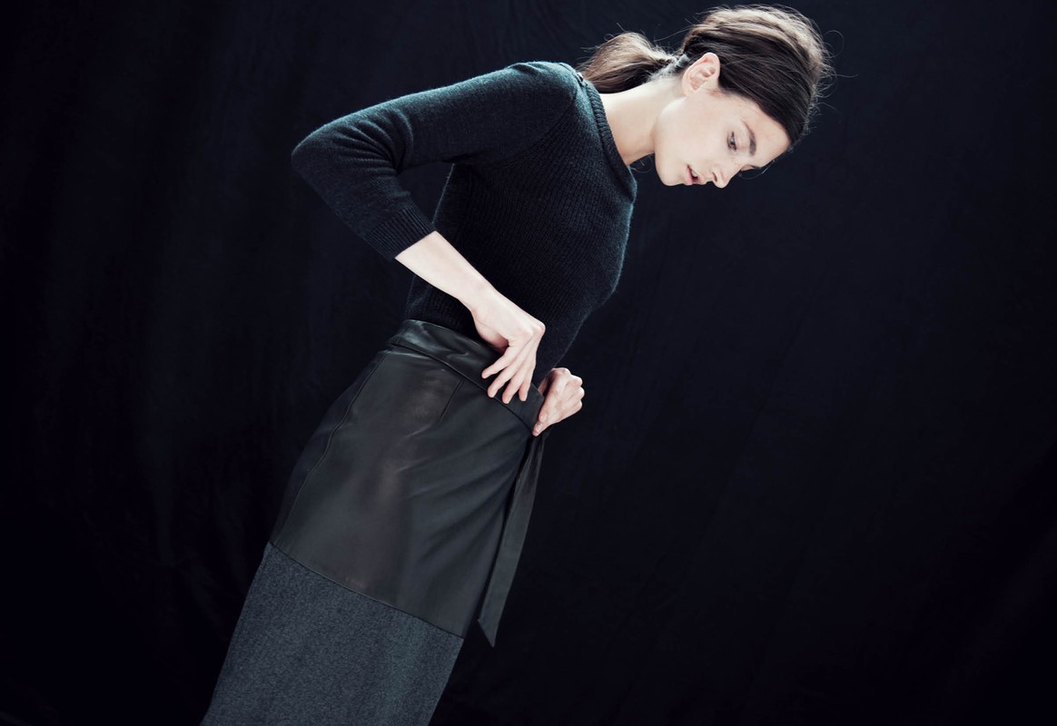 Woman in a fitted black sweater and wrap skirt poses in front of a dark background in a fashion style studio shot.