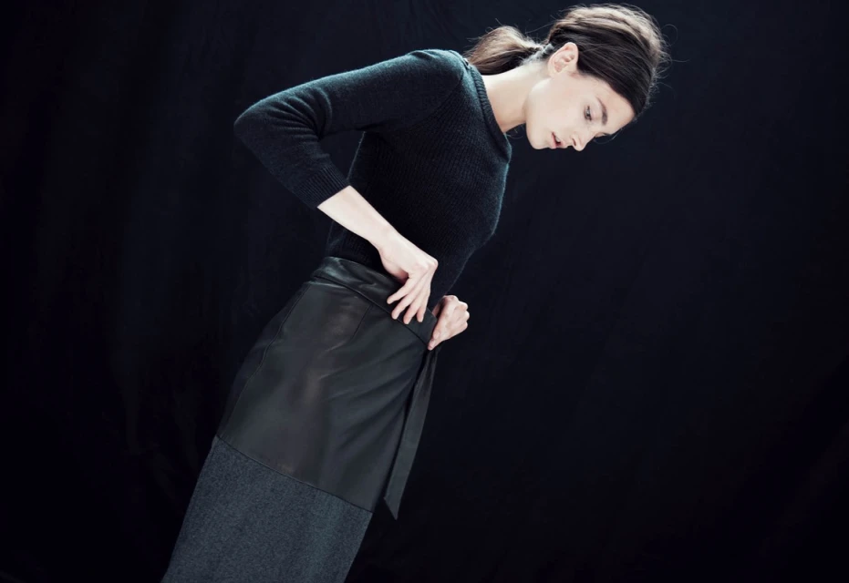 Woman in a fitted black sweater and wrap skirt poses in front of a dark background in a fashion style studio shot.