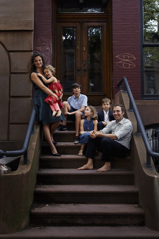Family portrait on brownstone steps, featuring a mother, father, and four children in an urban setting with warm, natural lighting.