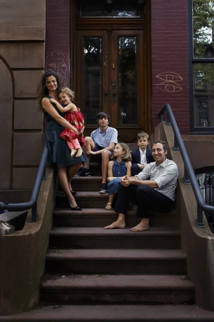Family portrait on brownstone steps, featuring a mother, father, and four children in an urban setting with warm, natural lighting.