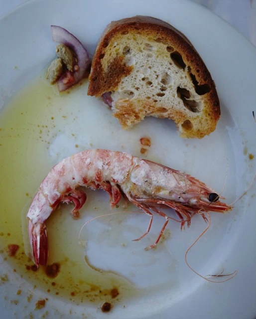 Overhead shot of cooked prawn alongside a piece of bread and a slice of onion on a white plate, with residual oil.