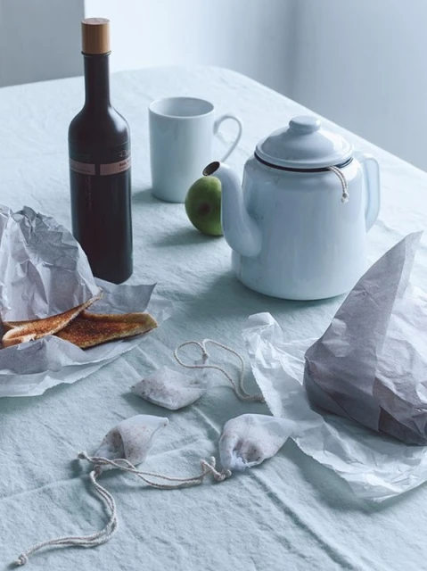 Still life photo of a bottle, apple, teapot, tea bags, mug, and food on a linen tablecloth, creating a serene and inviting composition.
