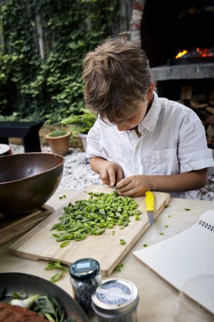 Young boy in a white shirt meticulously chops green beans on a wooden cutting board in an outdoor setting, surrounded by culinary ingredients.