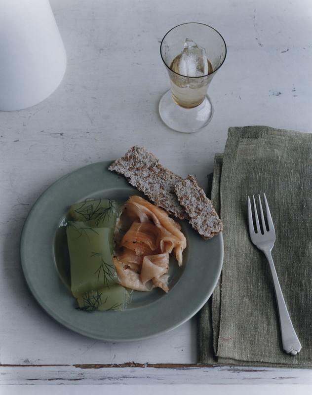 Overhead shot of a green plate with dill cucumbers, smoked salmon, crackers, a water glass, and a fork on a white wooden table with a green napkin.