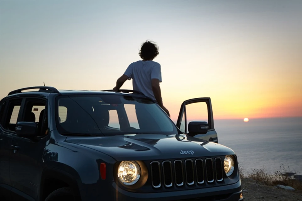 Dark grey Jeep on a hillside with a man standing on the roof, facing the ocean, sunset in the background, door open, outdoor setting.