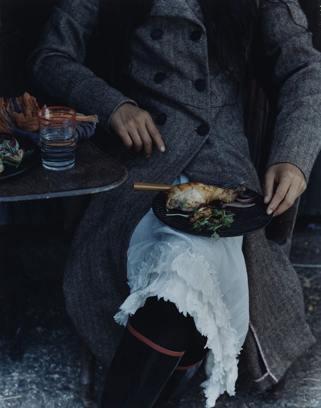 Person in tweed coat sits with plate of roasted chicken on lap and glass of water on table, creating a sophisticated food-focused image.