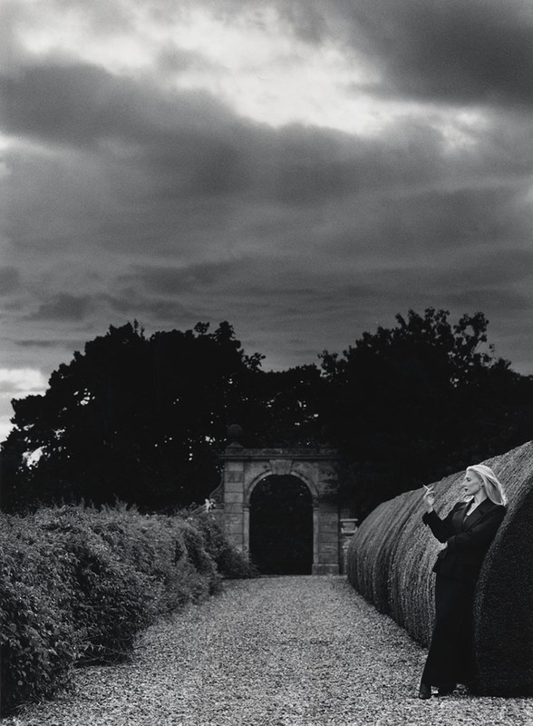 Monochrome outdoor shot of an adult woman in a suit, smoking, leaning against a sculpted hedge, with an archway and cloudy sky.