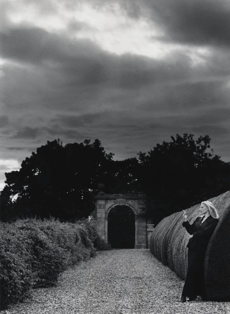Monochrome outdoor shot of an adult woman in a suit, smoking, leaning against a sculpted hedge, with an archway and cloudy sky.