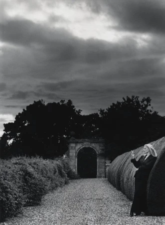 Monochrome outdoor shot of an adult woman in a suit, smoking, leaning against a sculpted hedge, with an archway and cloudy sky.