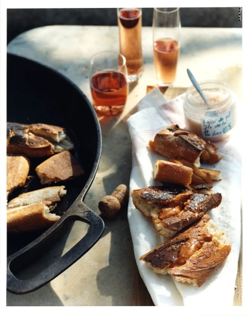 Toasted baguette slices sit on a table alongside glasses of rosé and a jar of flavored salt, capturing an inviting food still life.