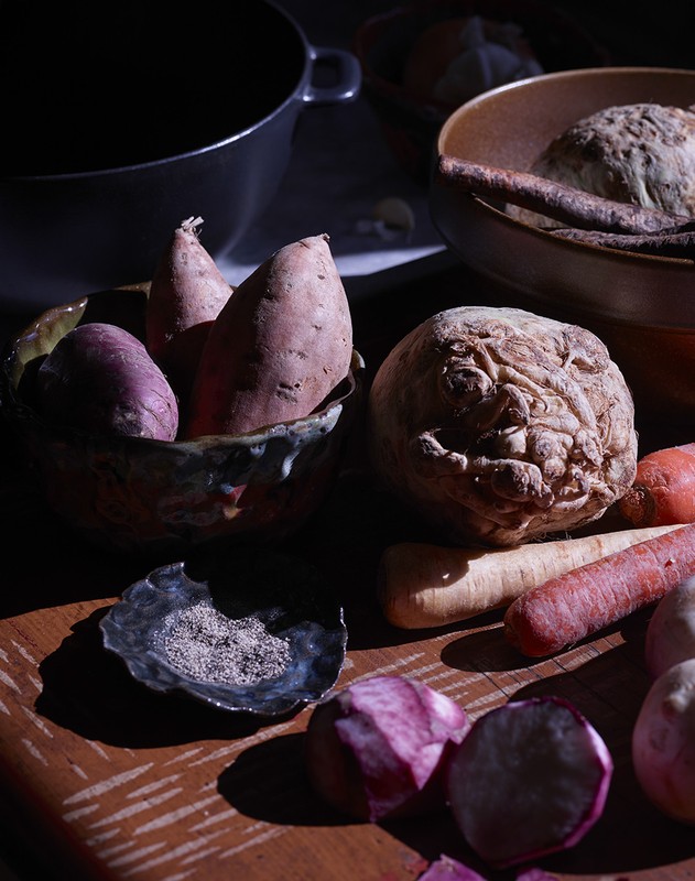 Still life shot of root vegetables including sweet potatoes, carrots, turnips, and celery root in rustic bowls on a wooden table.
