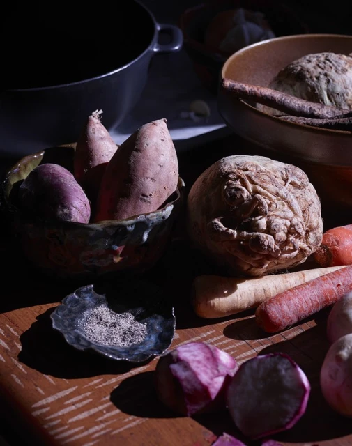 Still life shot of root vegetables including sweet potatoes, carrots, turnips, and celery root in rustic bowls on a wooden table.