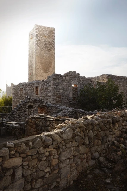 Stone ruins in rural setting under bright sky. An old, tall stone tower stands amid the weathered, crumbling stone architecture.
