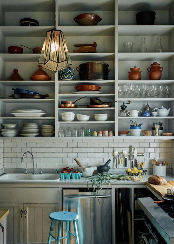 Bright kitchen interior with white shelves displaying pottery, glassware, and dishware, and featuring a blue stool and stainless steel dishwasher.