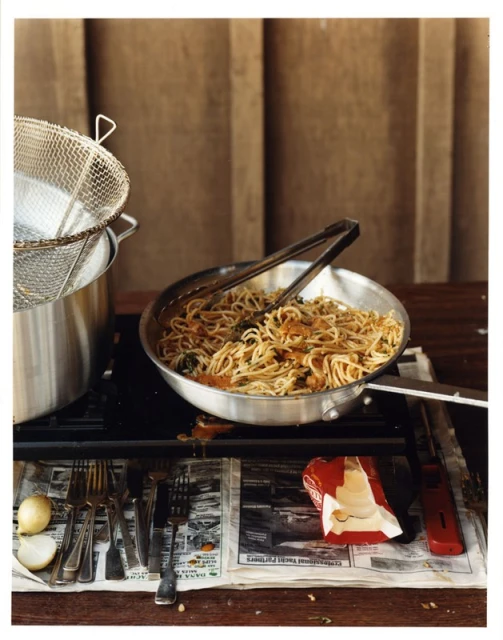 Overhead shot of spaghetti with tomato sauce in a metal pan on a stove, surrounded by kitchen utensils and an open newspaper.