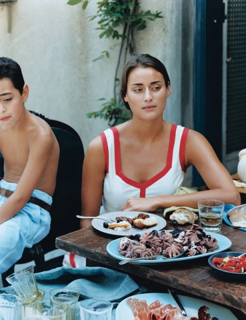 Woman and child sit at a table outdoors with dishes of food, including octopus, bread, and sliced meats, next to glassware and a blue towel.