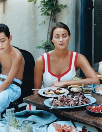 Woman and child sit at a table outdoors with dishes of food, including octopus, bread, and sliced meats, next to glassware and a blue towel.