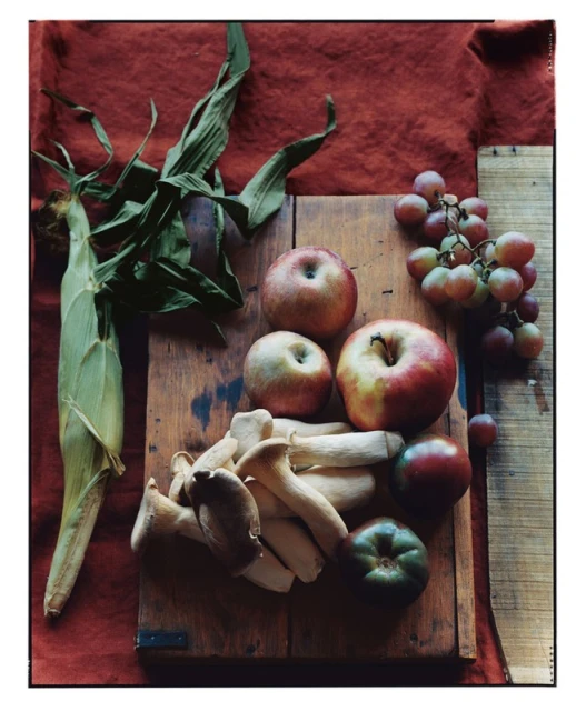 Still life photograph featuring a wooden board with apples, mushrooms, grapes, tomato, and corn, set against a textured fabric backdrop.