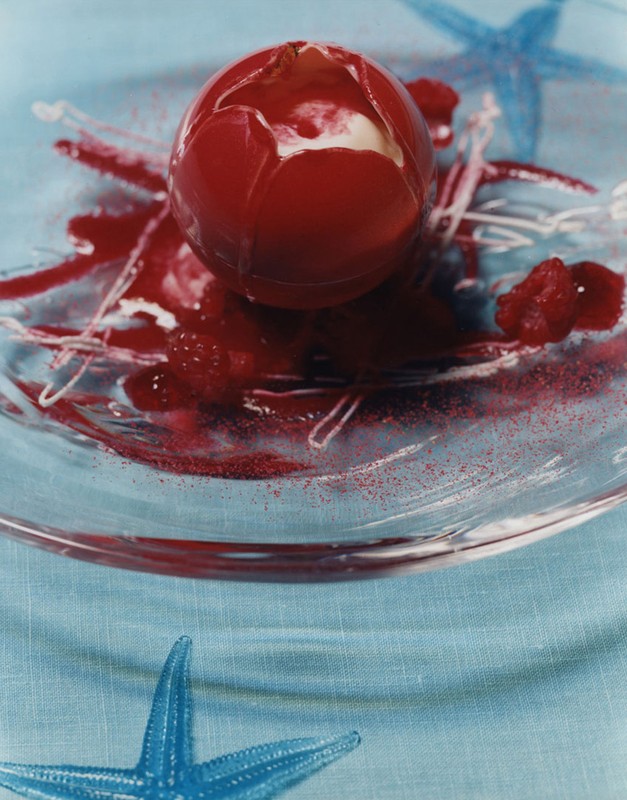 High-angle studio shot of a red dessert sphere on a clear plate with gel decorations, and blue starfish on a light blue tablecloth.