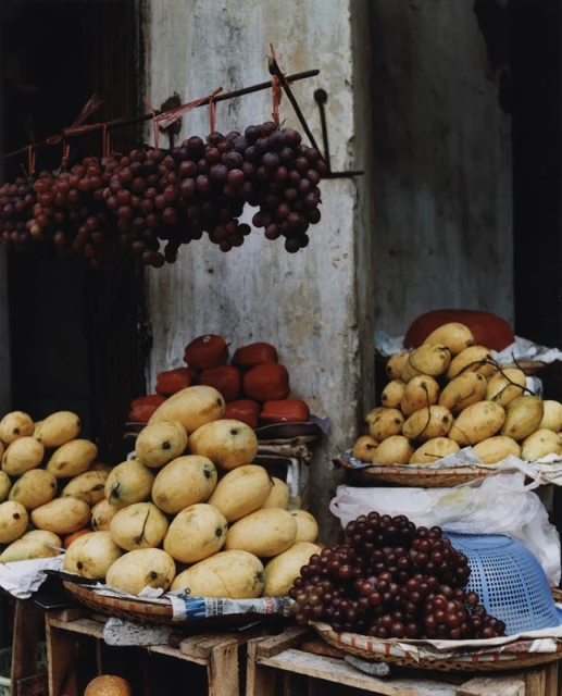 Outdoor produce stand with grapes hanging above mangos, red tomatoes, and blue baskets, creating a vibrant still-life scene.