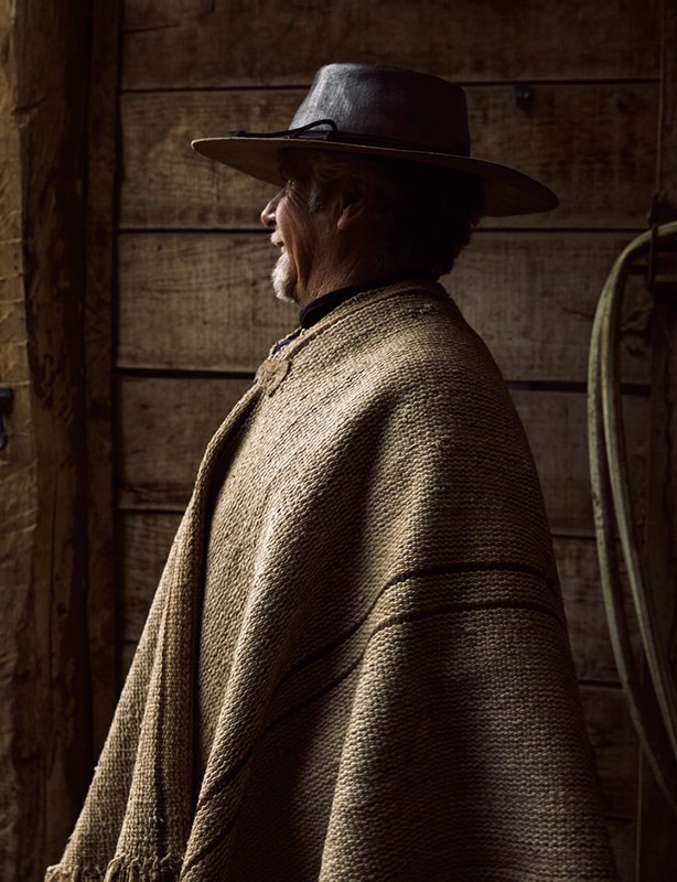 Profile shot of an elderly man wearing a poncho and hat, standing against a wooden wall with neutral tones and soft, diffused lighting.
