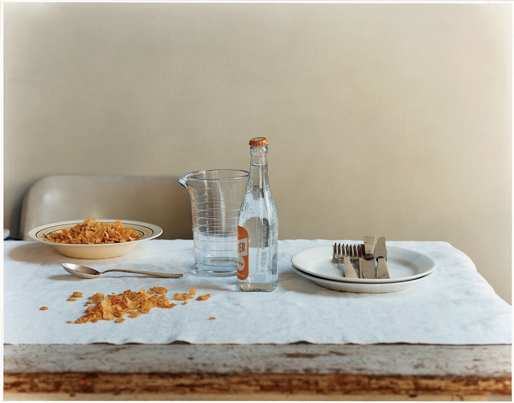 Still life photography of a breakfast table with a bowl of cereal, a bottle of water, and silverware on a white tablecloth indoors.