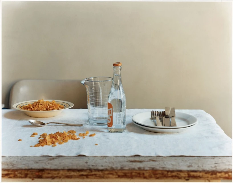 Still life photography of a breakfast table with a bowl of cereal, a bottle of water, and silverware on a white tablecloth indoors.