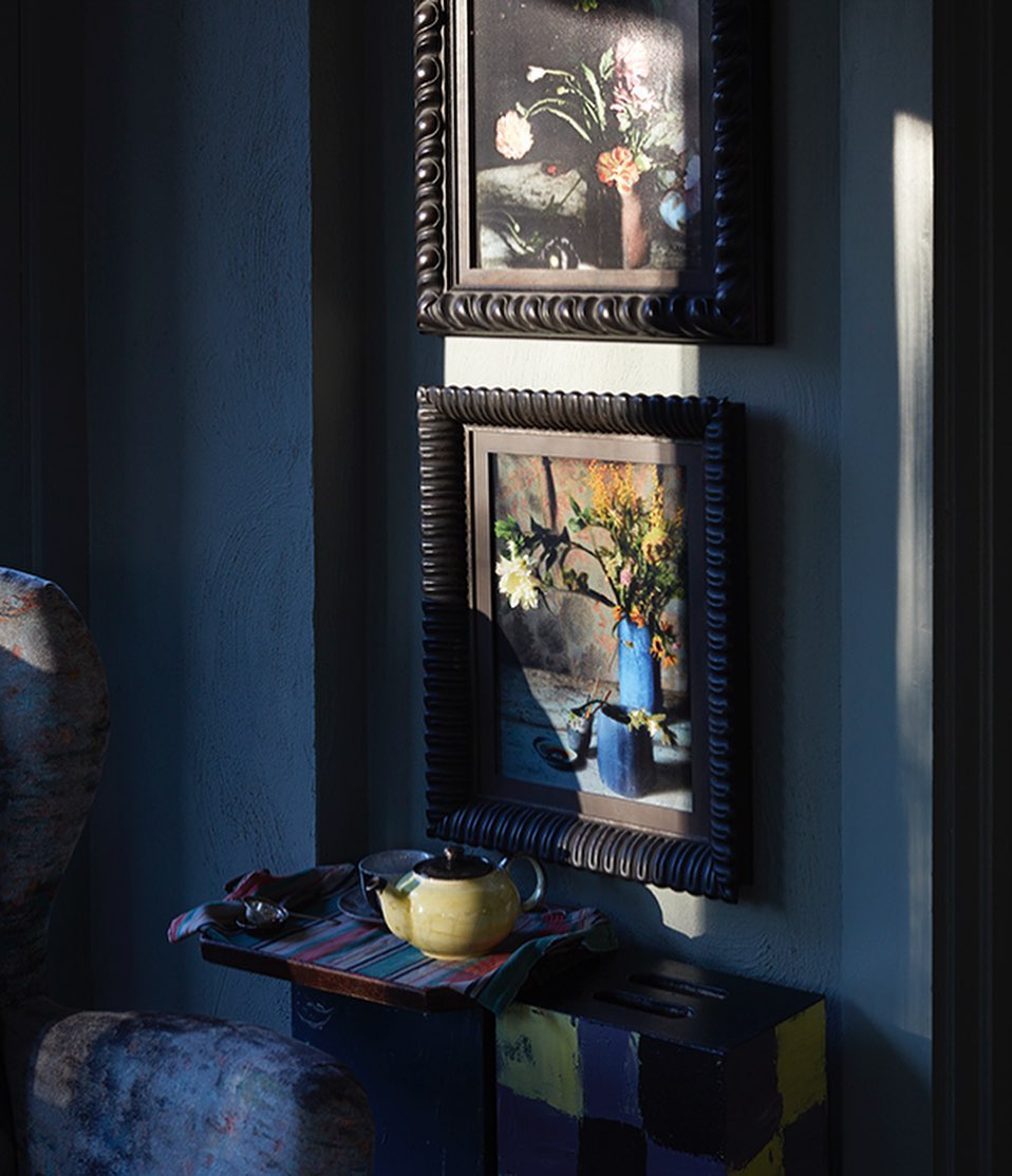 Two framed still-life paintings hang on a grey wall above a small table holding a tea set in a dimly lit room, creating a subdued ambiance.
