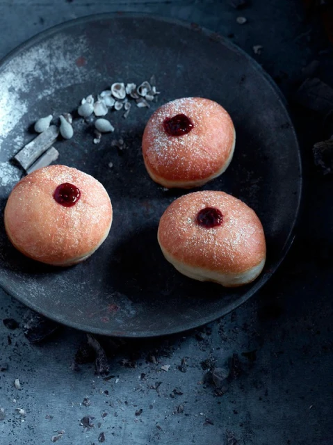 Three jam-filled doughnuts dusted with powdered sugar sit on a textured dark metal surface with nut pieces scattered around them.