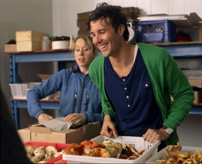 Two adults pack mushrooms into boxes at a table with wire shelving and other packed boxes visible in the background, inside.