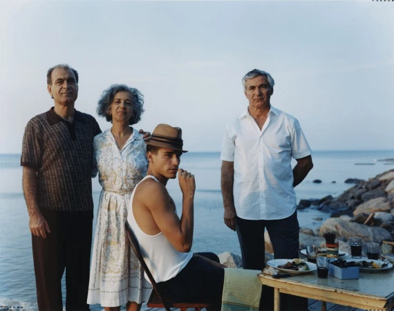 Four people pose outdoors by the sea at a table with drinks and food, in a medium shot with neutral light and natural background.