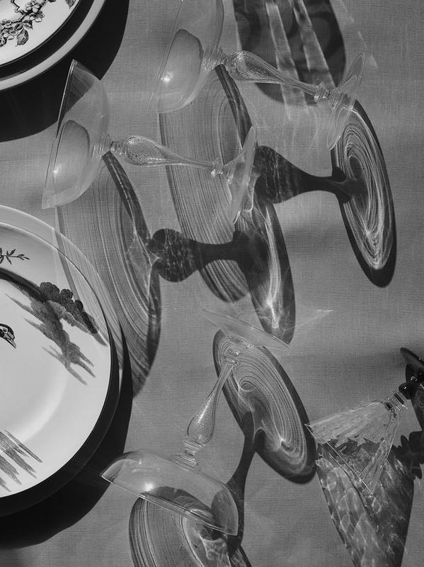 Monochrome still life of wine glasses and decorative plates with shadows creating abstract patterns on textured surface.